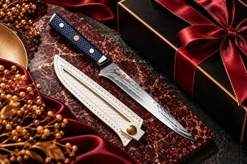 Damascus chef’s knife with navy handle and ivory sheath on burgundy marble, styled with a black gift box and gold accents.