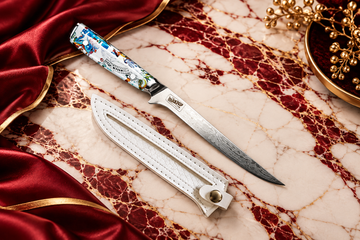 Premium Damascus kitchen knife with a colourful handle and white leather sheath on burgundy red and white marble with elegant gold accents.