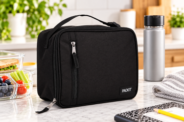 Black PackIt freezable lunch bag on a kitchen countertop with fresh food and a water bottle, illustrating lunch bag care and maintenance.